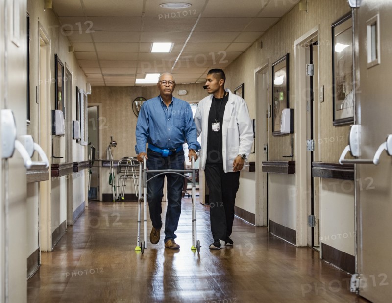 Male nurse assisting senior man walking down corridor with mobility walker