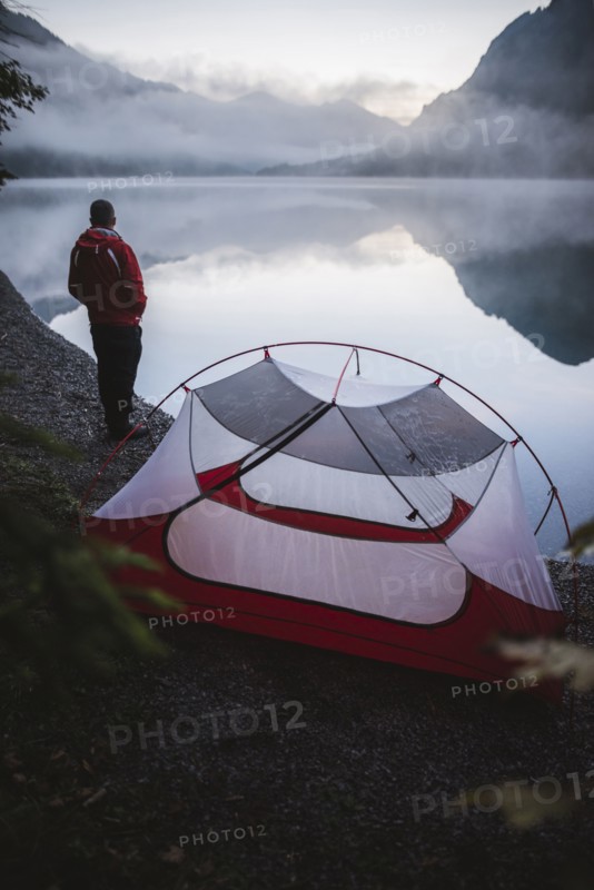 Austria, Plansee, Young man standing near tent and looking at Plansee lake at sunrise