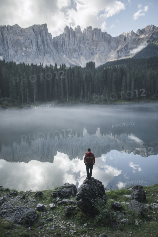 Italy, Carezza, Young man standing on rock at Lago di Carezza in Dolomite Alps at dawn