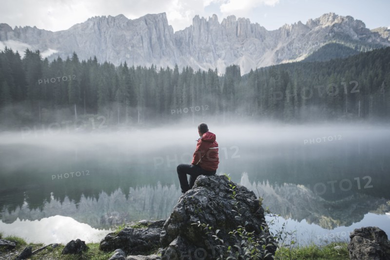 Switzerland, Young man sitting at Lago di Carezza in Dolomite Alps at dawn