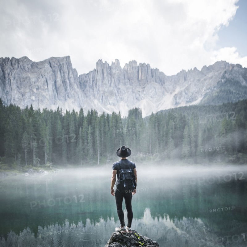 Italy, Carezza, Young man standing on rock at Lago di Carezza in Dolomite Alps at dawn
