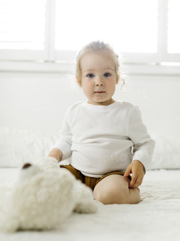 Smiling toddler girl sitting on bed with teddy bear toy