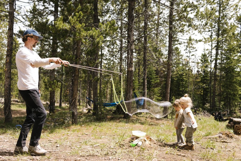 Father making large soap bubbles in forest for two daughters