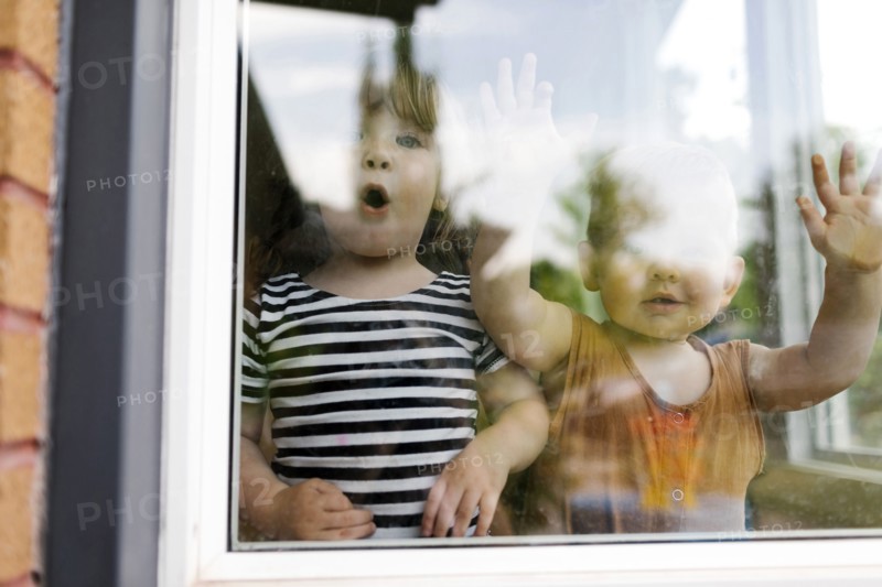 Portrait of toddler girl (2-3) and baby boy (18-23 months) standing behind window