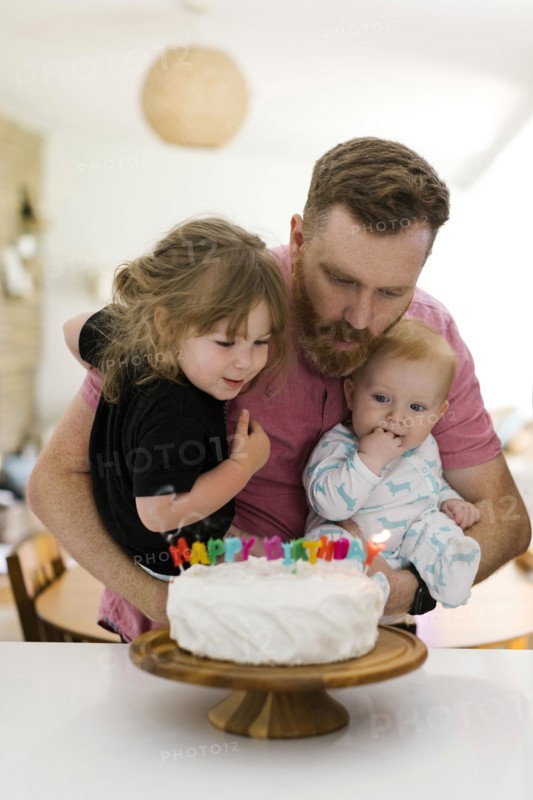 Father carrying kids (2-3, 6-11 months) and blowing birthday candles on cake