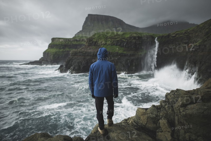 Denmark, Faroe Islands, Gasadalur village, Mulafossur Waterfall, Man standing on cliff and looking at Mulafossur Waterfall falling into ocean