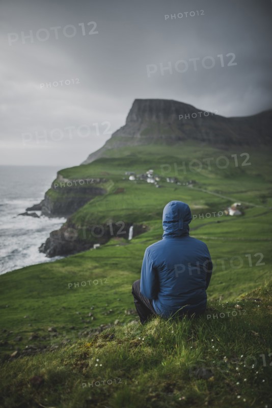 Denmark, Faroe Islands, Gasadalur village, Man sitting and looking at cliffed coast and ocean