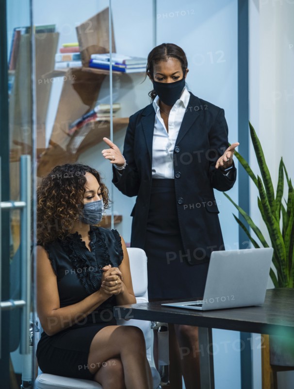 Two businesswomen wearing face masks working on laptop at desk in office