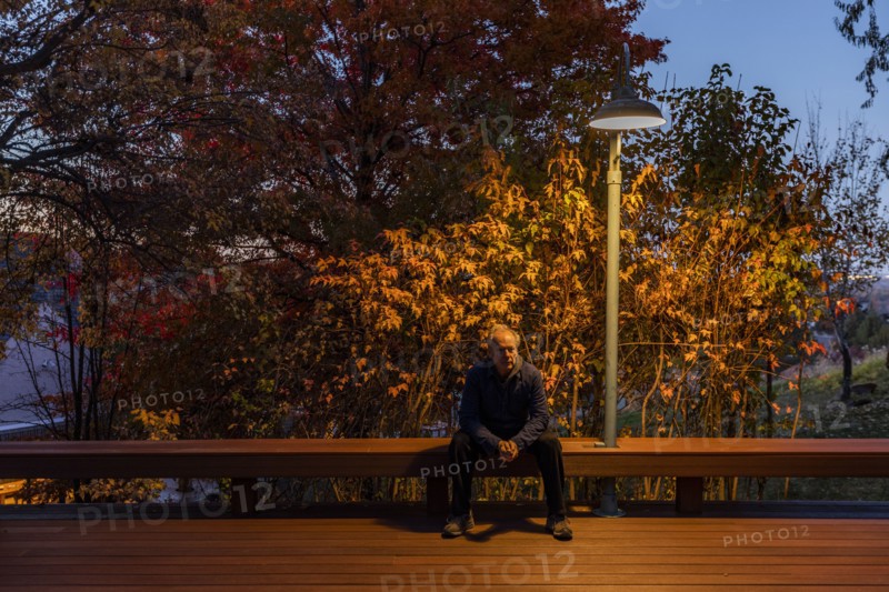 Senior man sitting on deck bench under autumn tree illuminated at night