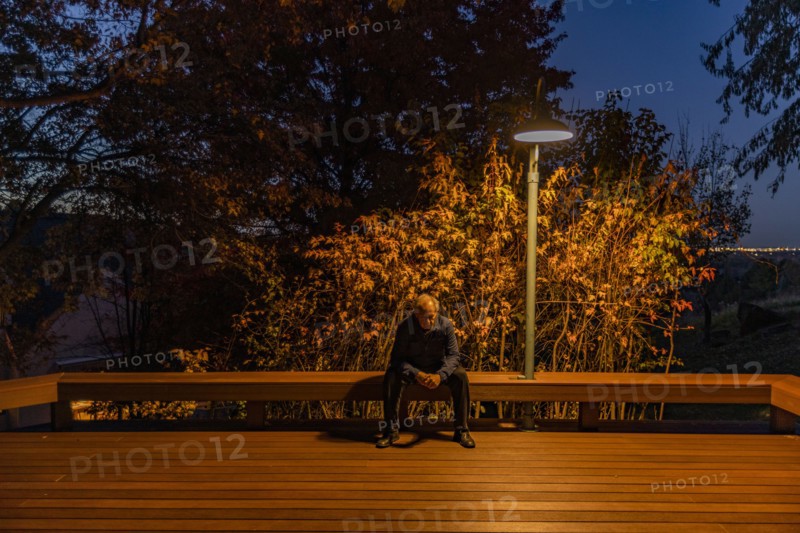 Senior man sitting on deck bench under autumn tree illuminated at night