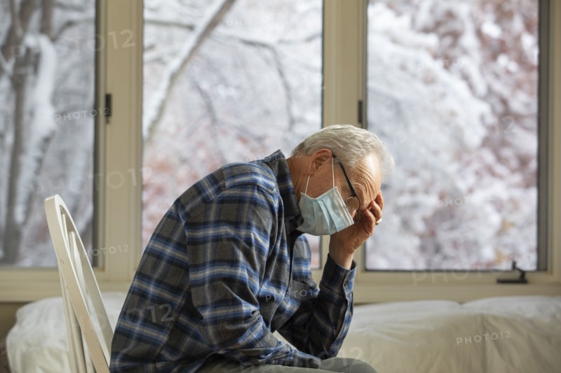 Senior man in Covid protective mask sitting on chair by window