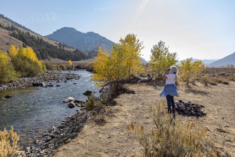 Senior woman looking through binoculars while standing by river in autumn landscape