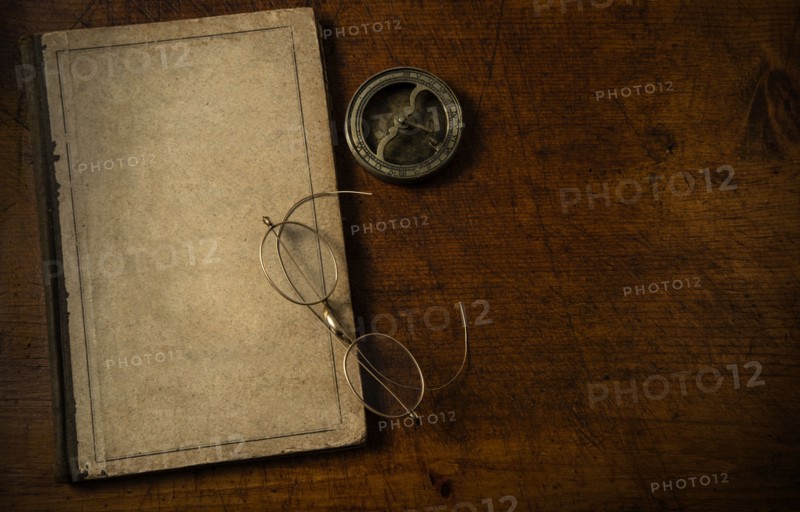 Old book with blank cover resting on old wooden desk top with old eyeglasses and small sundial