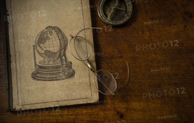 Old book resting on old wooden desk top with old eyeglasses and small sundial