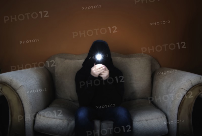 Man in hood sitting on sofa and holding flashlight in front of his face