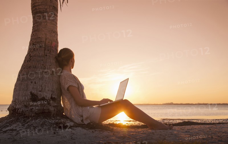 Caucasian woman with laptop leaning on palm tree at sunset