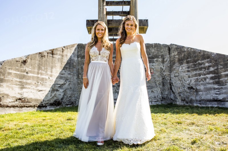 Caucasian brides holding hands in grass near concrete structure