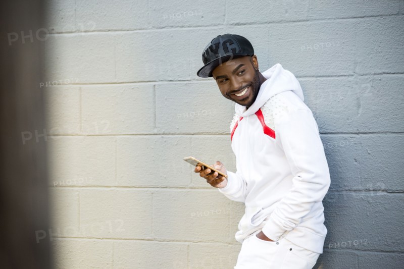 Smiling Black man leaning on wall holding cell phone