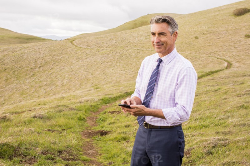 Smiling Caucasian businessman standing in grass holding cell phone