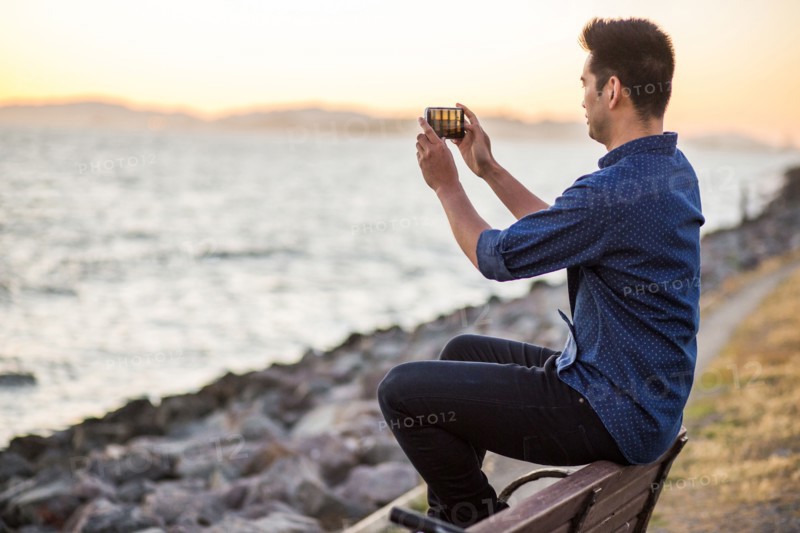 Chinese man sitting on bench photographing ocean with cell phone