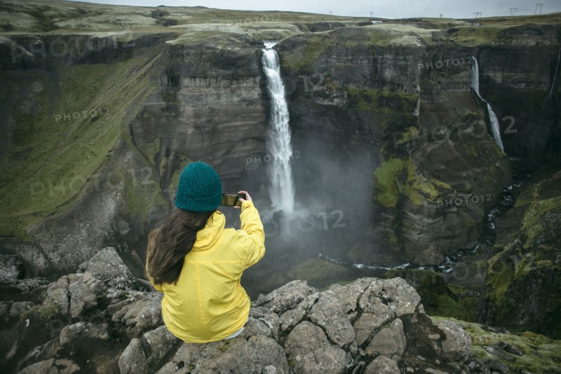 Caucasian woman sitting on cliff photographing waterfall
