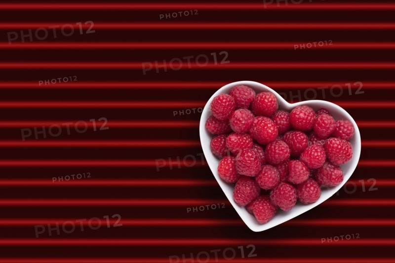 Raspberries in heart-shape bowl on red striped background