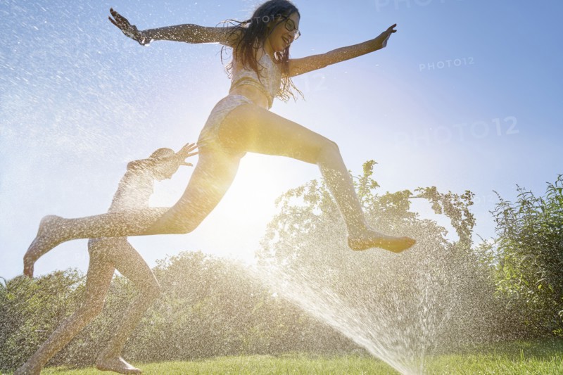 Caucasian girls running and jumping through backyard sprinkler