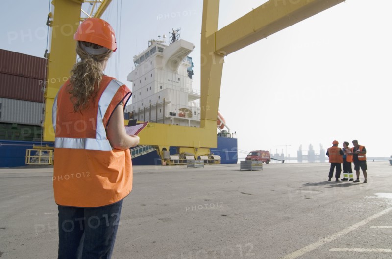 Woman wearing hard hat holding clipboard standing on dockside