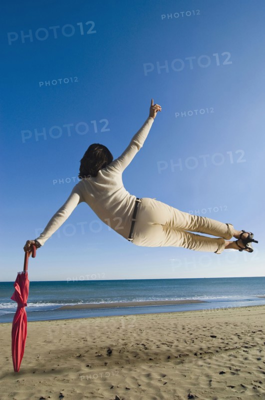 Woman jumping mid air on beach leaning on umbrella rear view