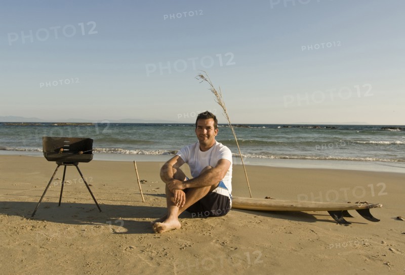 man sitting on beach with surfboard and barbecue