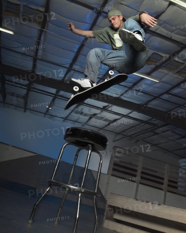 Caucasian man jumping on skateboard over stool indoors