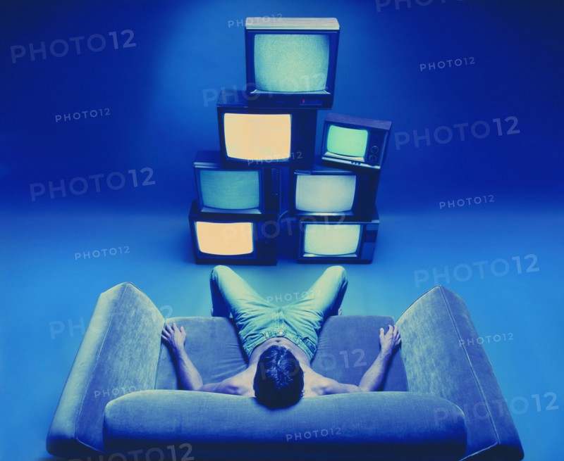 Man sitting on loveseat watching stack of televisions