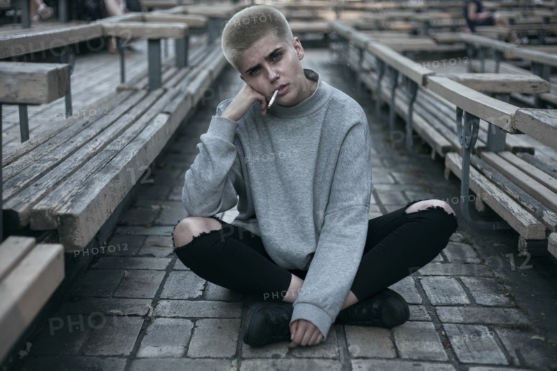Punk Caucasian woman sitting on cobblestones smoking cigarette
