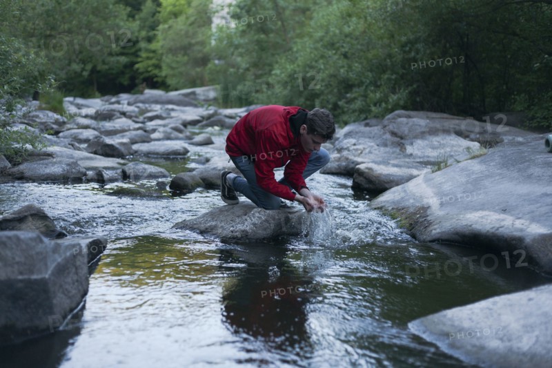 Caucasian man kneeling on rock in river cupping water with hands