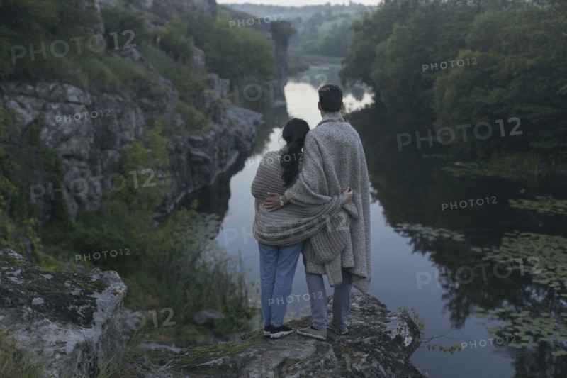 Caucasian couple wrapped in blanket standing on rock admiring river