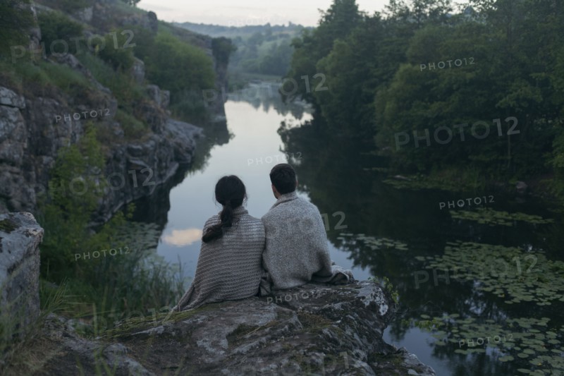 Caucasian couple wrapped in blanket sitting on rock admiring river