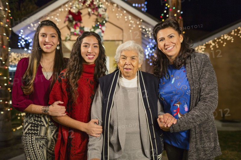 Three generations of Hispanic women smiling outside house decorated with string lights