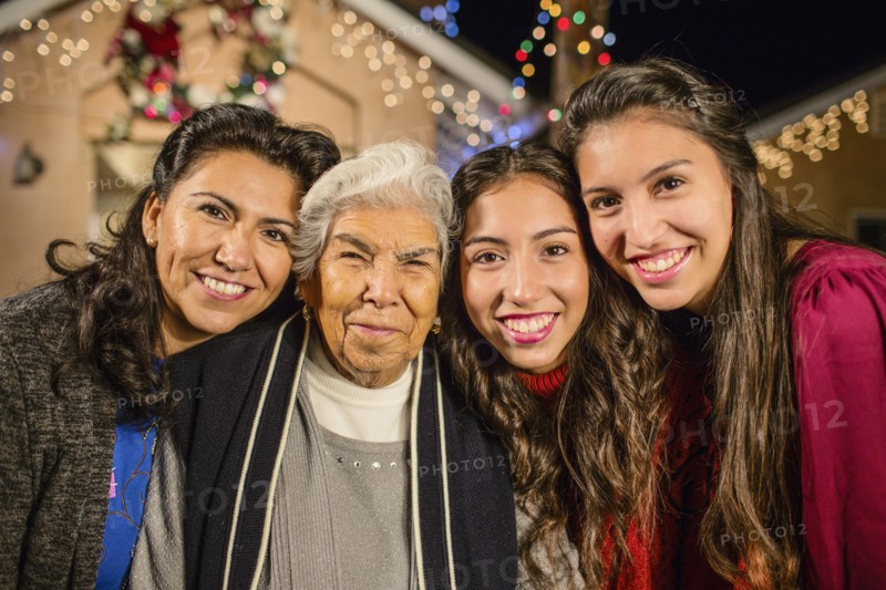 Three generations of Hispanic women standing outside house decorated with string lights