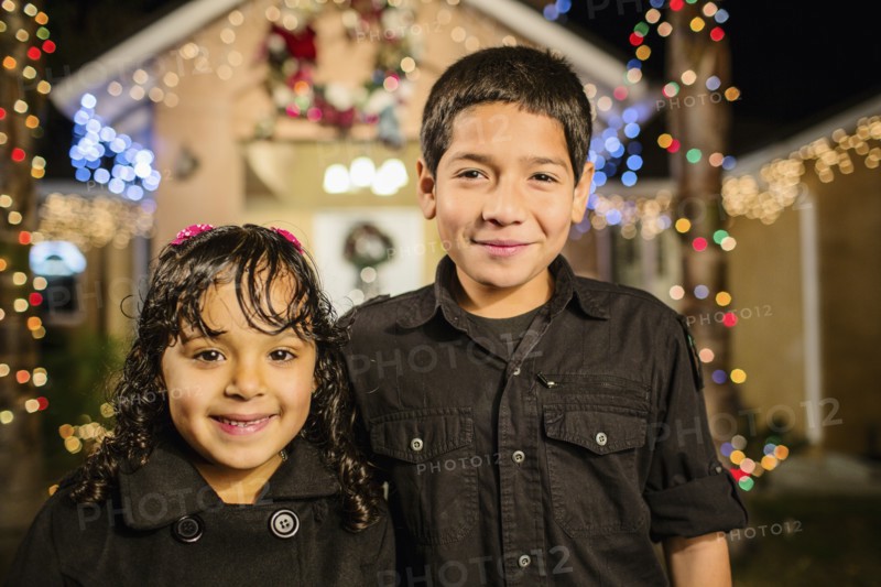 Hispanic brother and sister smiling outside house decorated with string lights