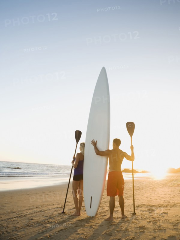 Couple standing on beach with surfboard and paddles