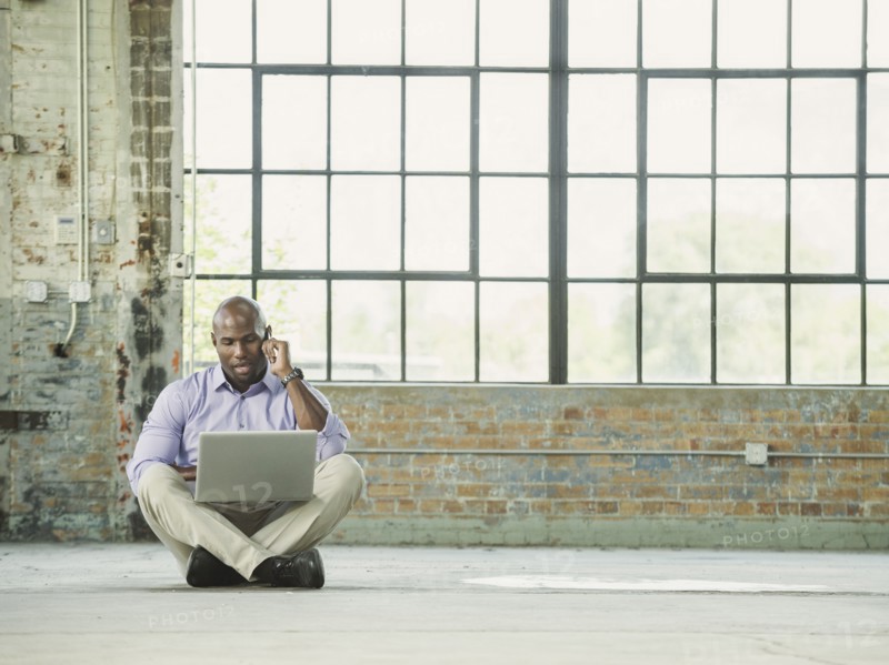 Black businessman using laptop and cell phone on warehouse floor