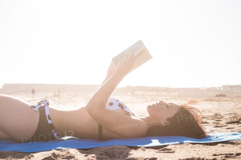 Caucasian woman laying on beach blanket reading book