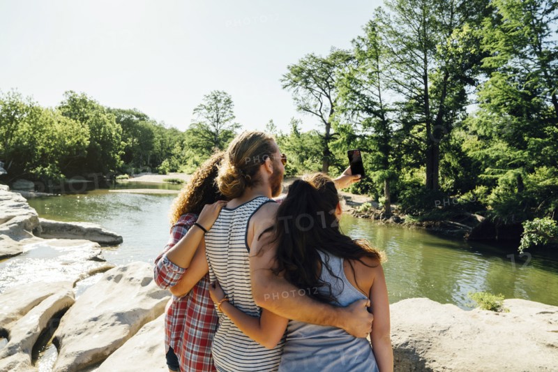 Friends posing for cell phone selfie on rocks near river