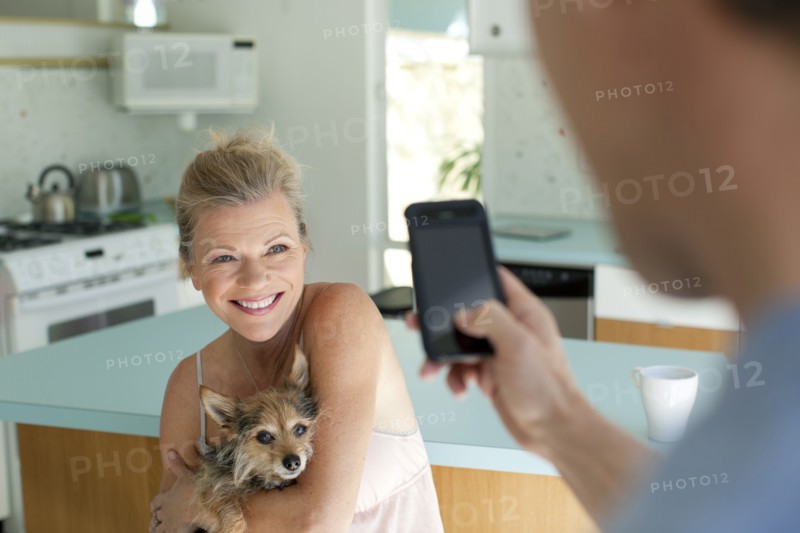 Man taking picture of wife holding dog in kitchen