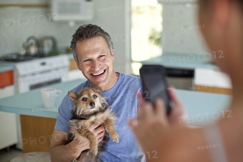 Woman taking picture of husband holding dog in kitchen