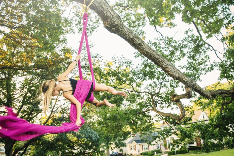Acrobatic Caucasian girl hanging on fabric under tree