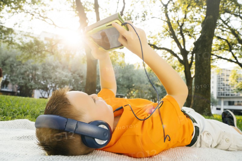 Caucasian boy laying on blanket in park listening to digital tablet