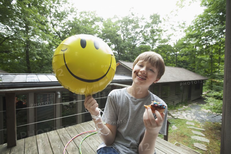 Caucasian boy with messy face holding smiley face balloon and eating donut