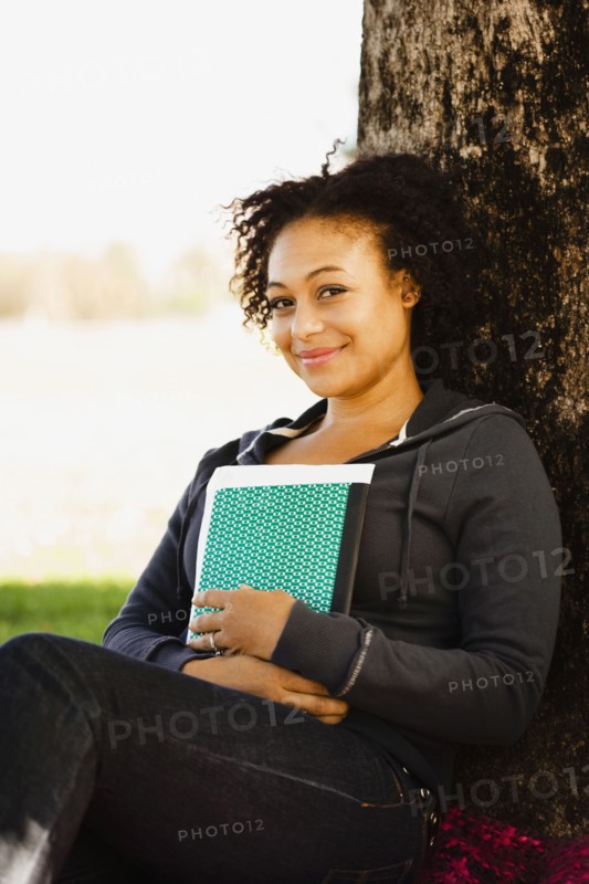 African woman holding book leaning against tree
