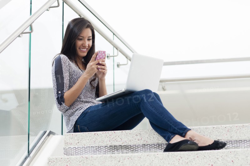 Hispanic woman using cell phone and laptop on staircase